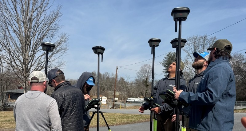 Surveyors participating in GNSS field training, using multiple rover poles with receivers attached while standing beside a roadside.
